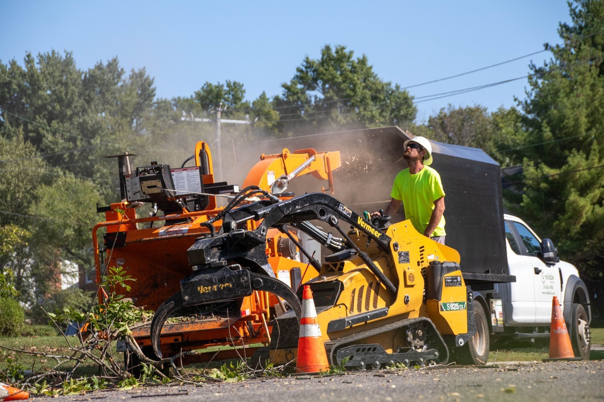 Worker feeding branches into a wood chipper; small excavator beside a truck on a sunny day.