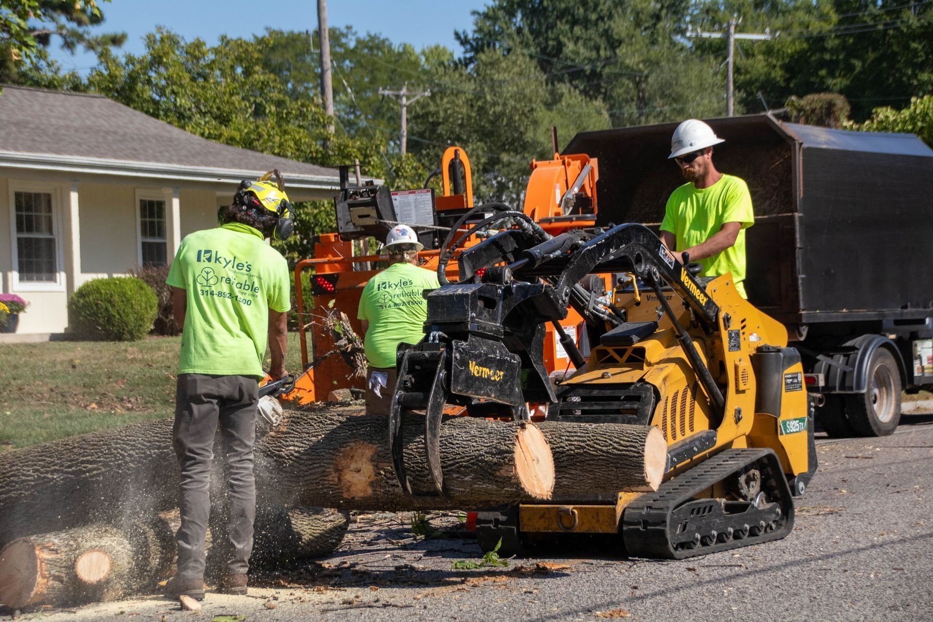 Tree removal crew operating machinery on a street. Workers in safety gear, yellow loader and truck.