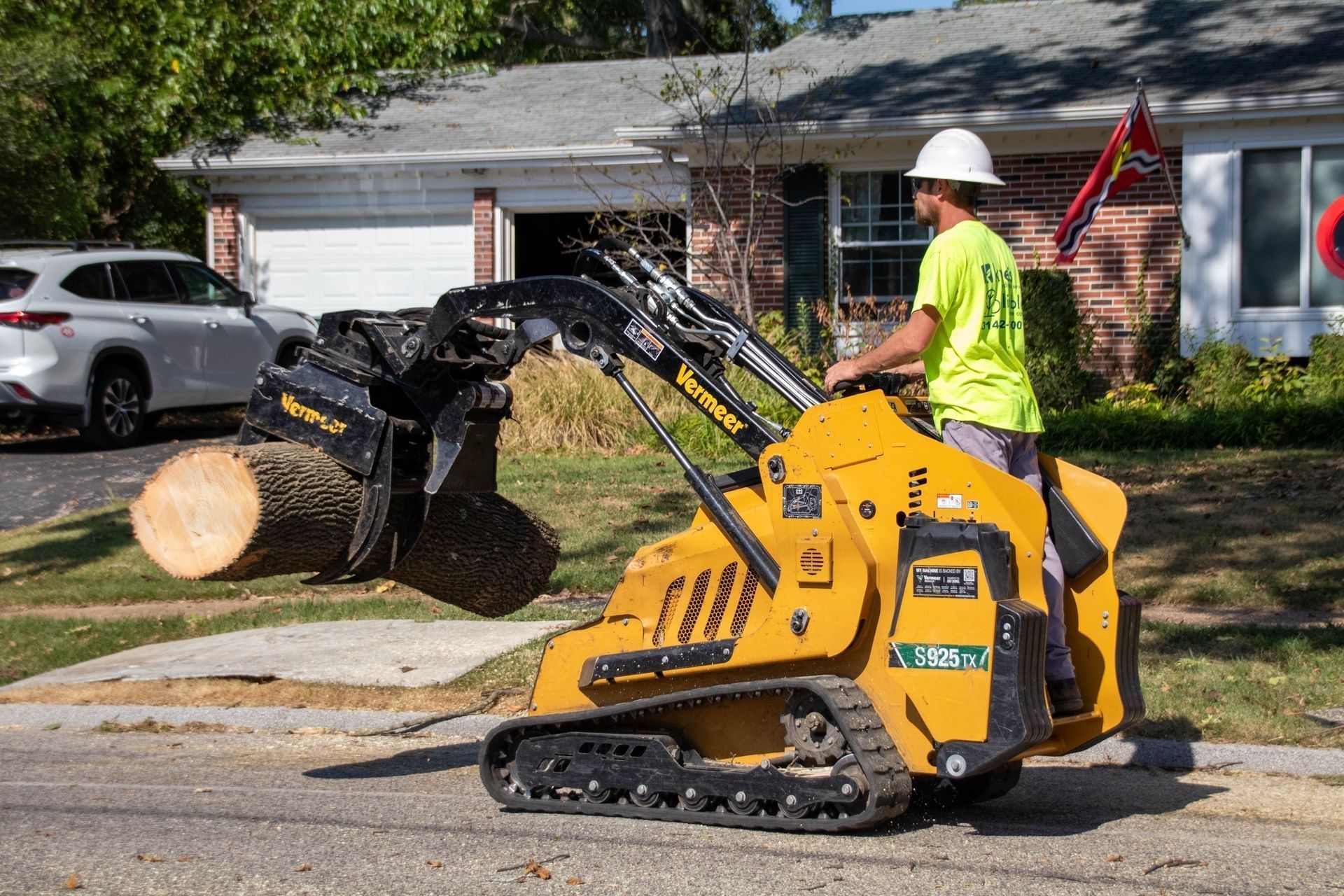 Man operating a yellow skid steer, lifting a log on a suburban street.