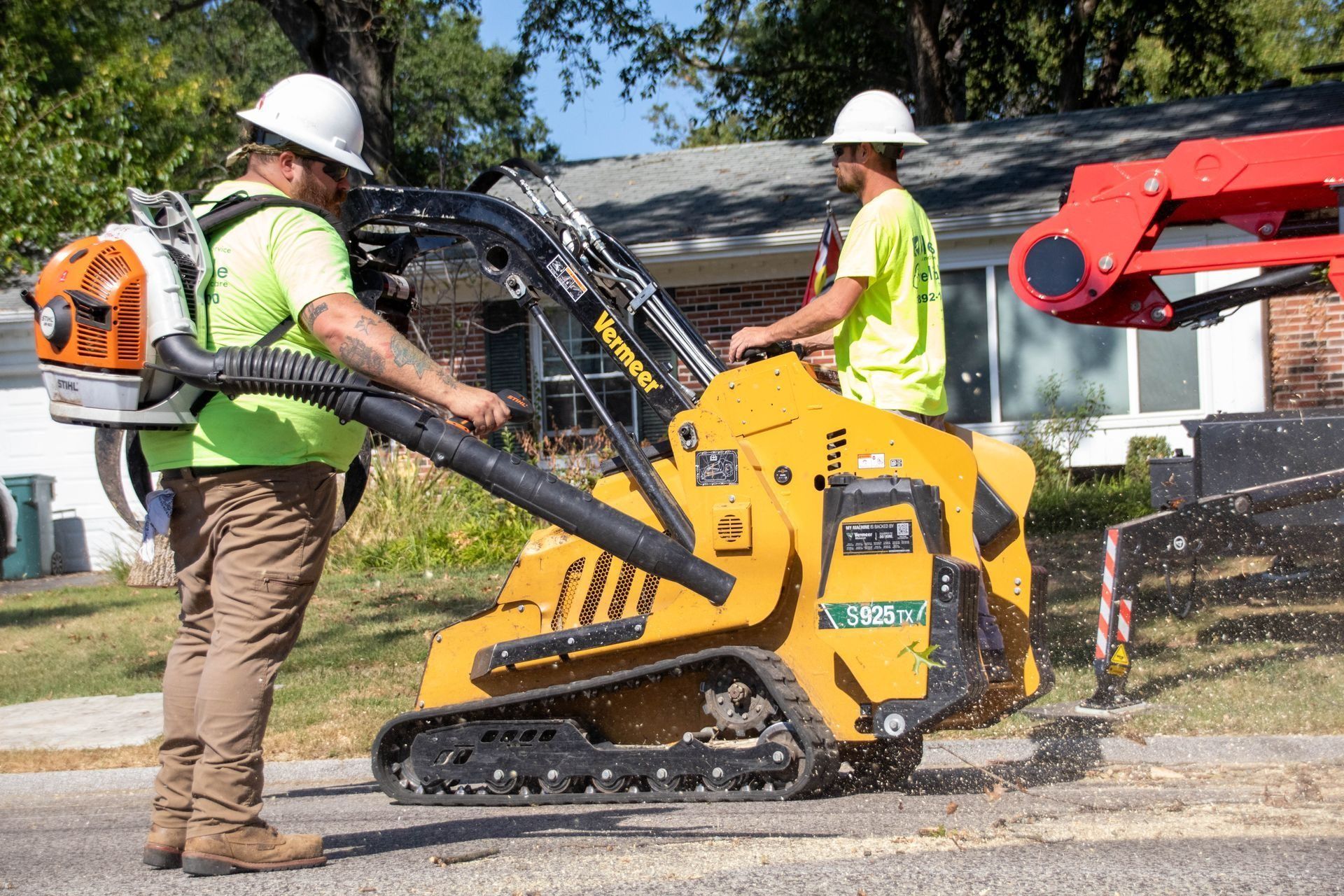 Two workers using equipment on a street. One uses a leaf blower, the other operates a tracked mini-skid steer.
