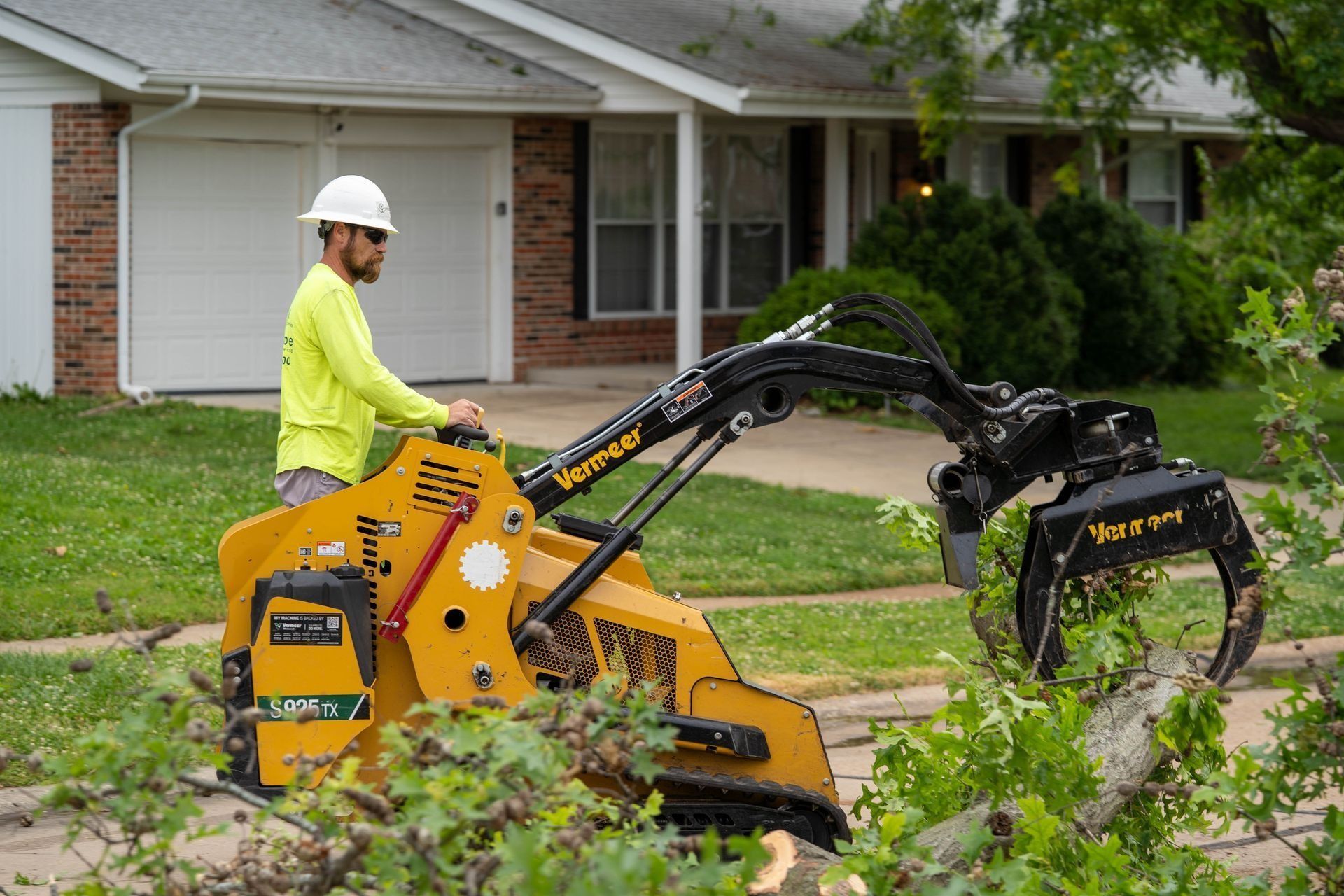 Worker operating a yellow tree-cutting machine in front of a house.