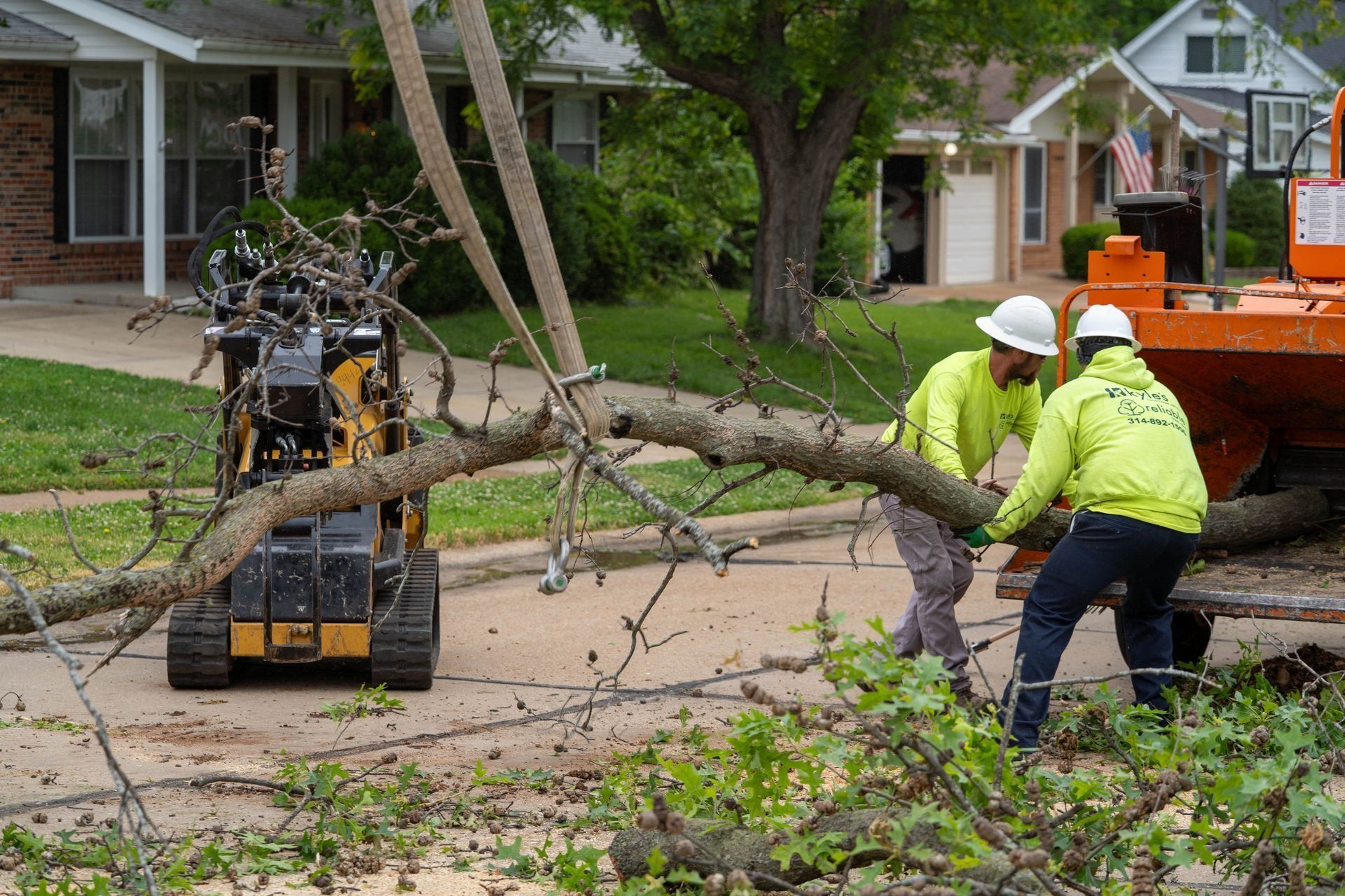 Two workers in neon vests lift a tree branch while a skid steer holds it in a residential street.