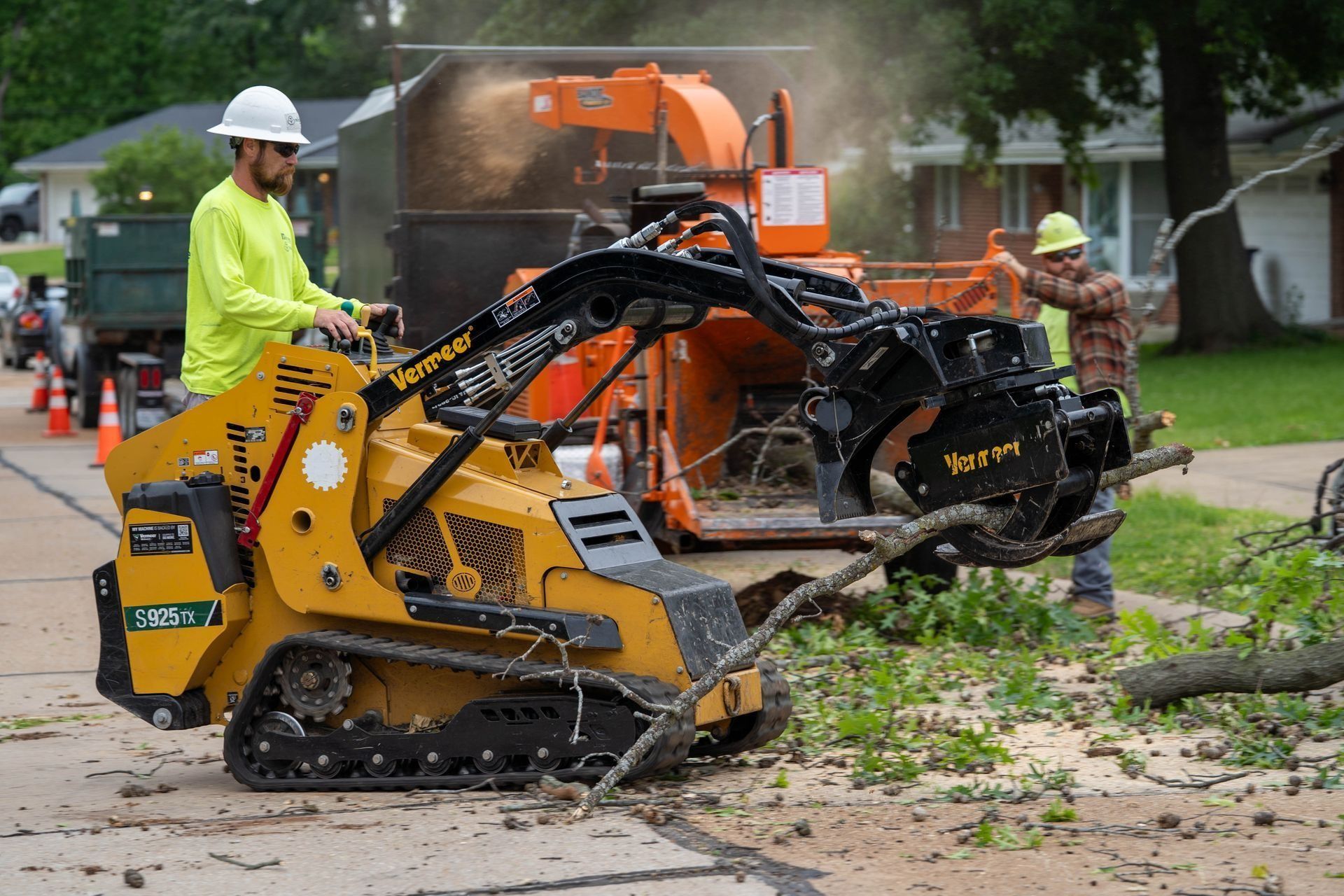 Workers using a skid steer with grapple and wood chipper to clear branches from a street.