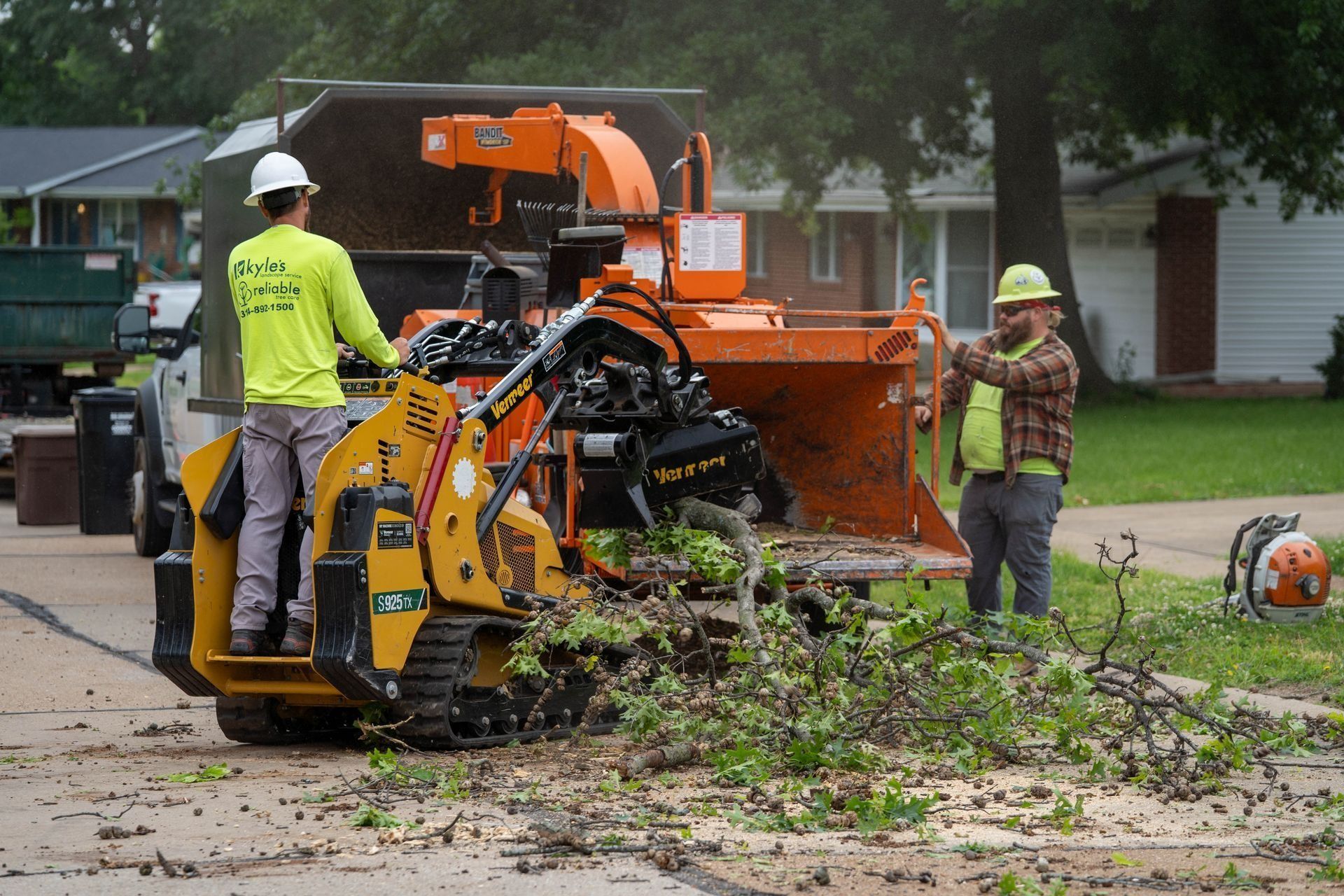Two workers chipping tree branches on a street with an orange chipper; one drives the machine, the other feeds it branches.