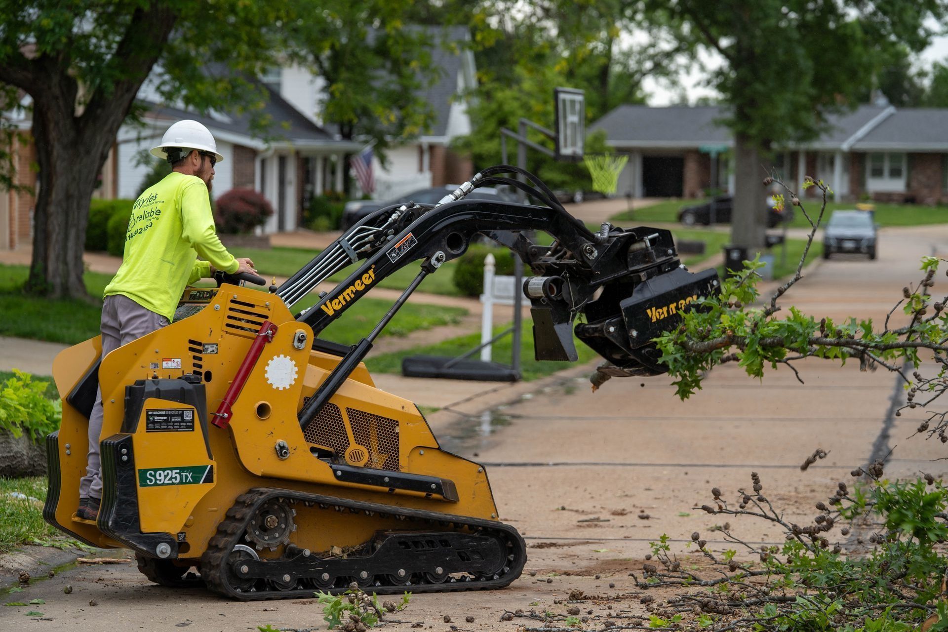 Yellow skid steer trimming tree branches on a street with a worker wearing a hard hat and neon shirt.