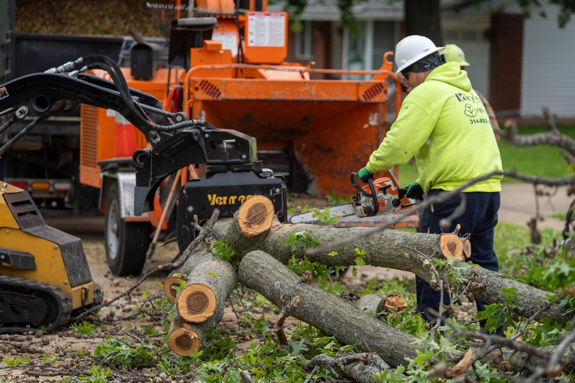 Man in a neon jacket uses a chainsaw to cut tree limbs near an orange wood chipper and skid steer.