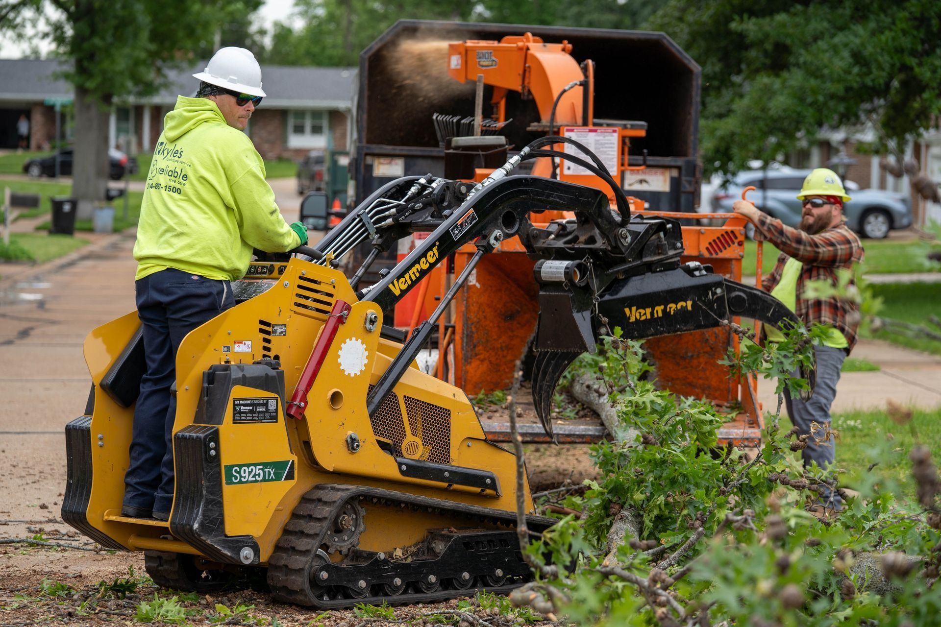 Workers using a skid steer with grapple and a wood chipper to clear tree branches in a neighborhood.