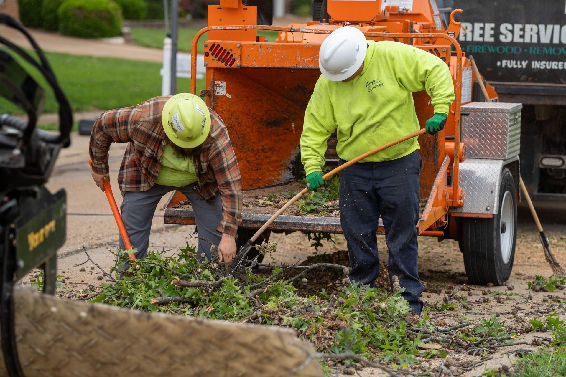 Two workers in safety gear loading brush into a wood chipper on a street.