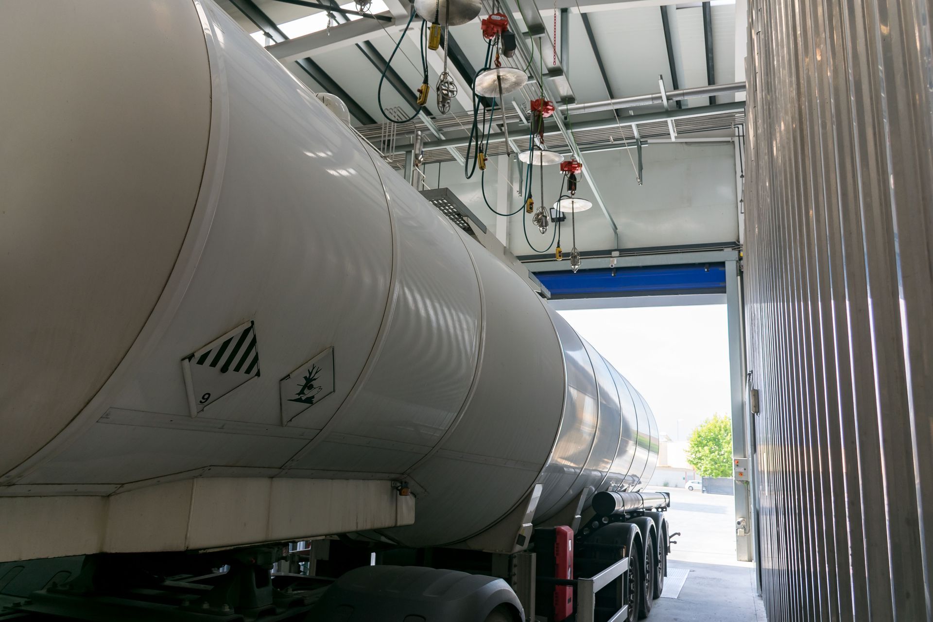 Tanker truck parked at a loading dock, under an overhead structure with piping and connectors.
