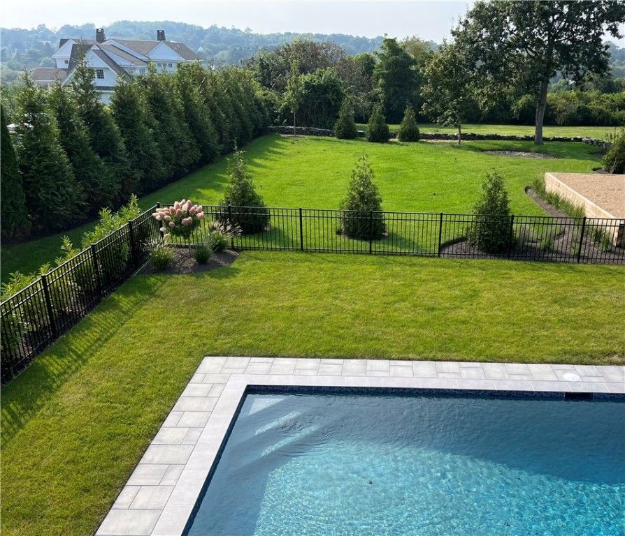 Backyard with pool, lawn, and evergreen trees behind a black metal fence, under a blue sky.