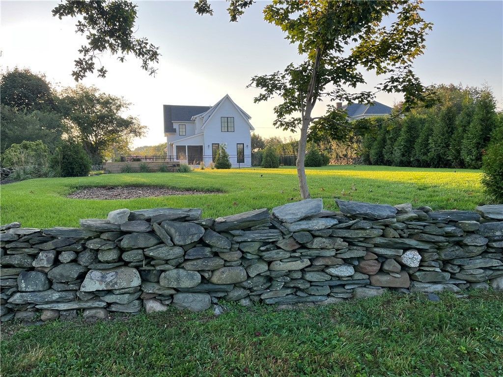White house with black roof, set beyond stone wall and green lawn, under a clear sky.