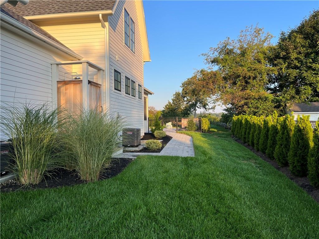 Side of a white house with manicured lawn, stone pathway, and line of green trees on a sunny day.