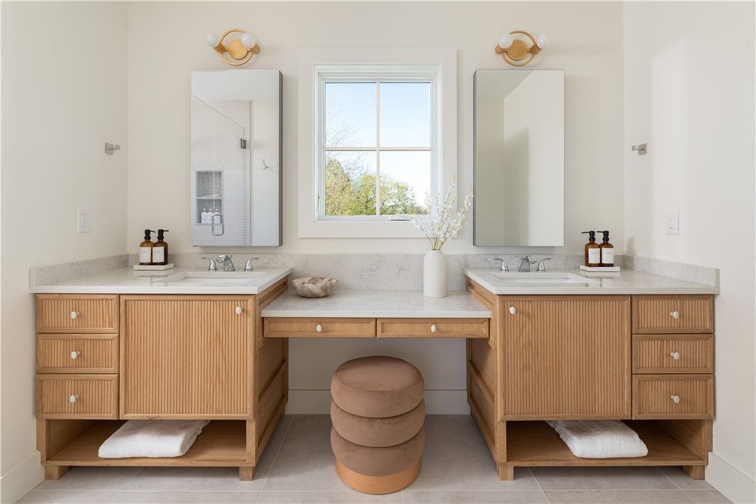 A modern bathroom with two wood vanities, a center stool, and a window.
