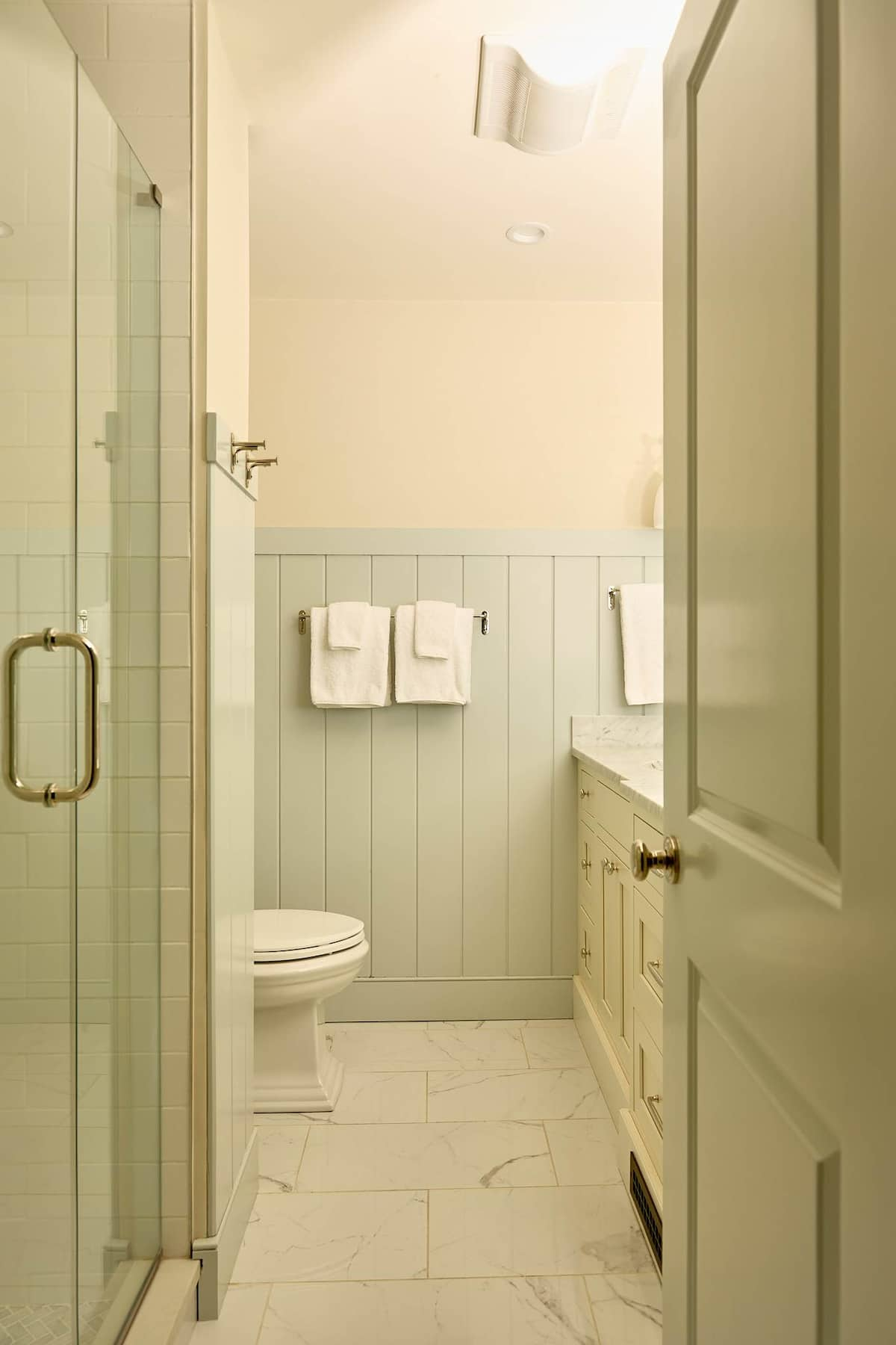 Narrow bathroom with a glass shower, toilet, light-blue wainscoting, and white vanity.