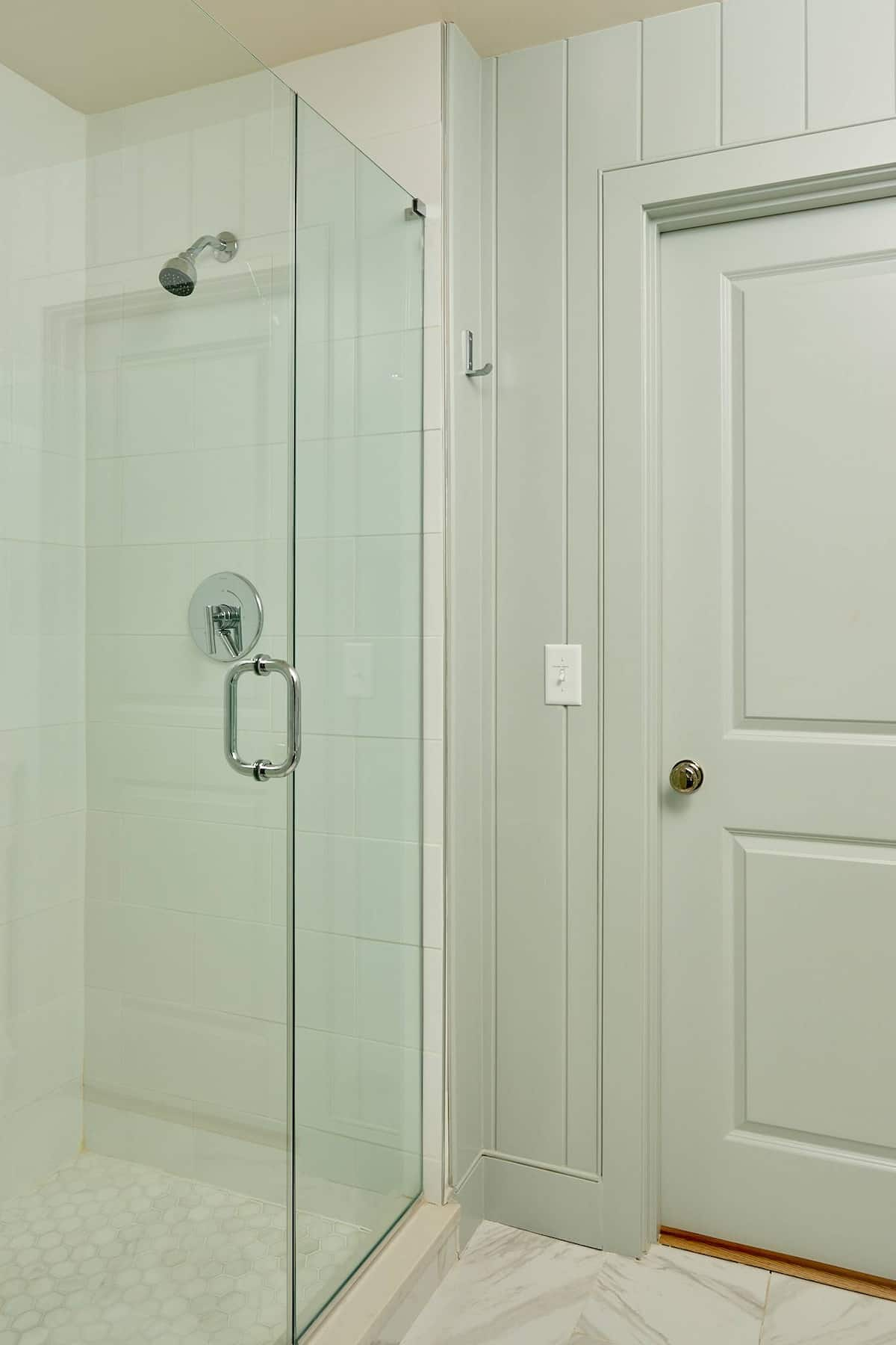 Shower with glass door next to a gray door. White walls, chrome hardware.