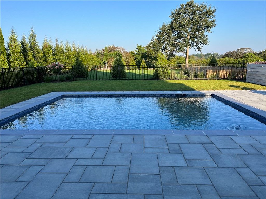 Rectangular pool surrounded by blue pavers and lawn. Trees and fence in background under a sunny sky.