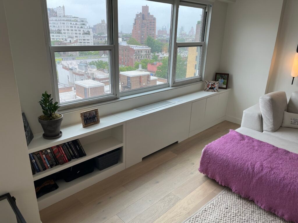 Bedroom with window, built-in cabinets, and a sofa covered with a purple blanket. View of city buildings visible.