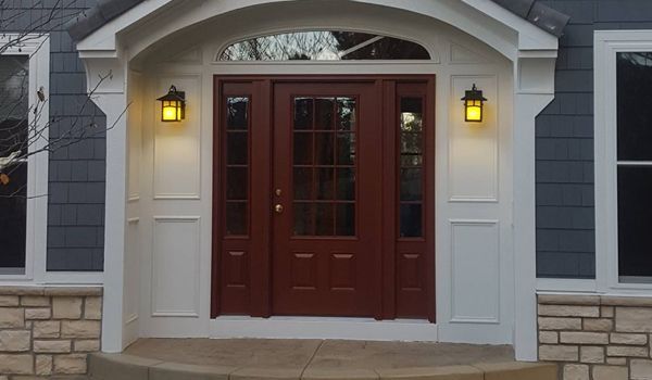 Wooden front door with glass panels, sidelights, and overhead transom, flanked by white pillars and sconces; blue siding.