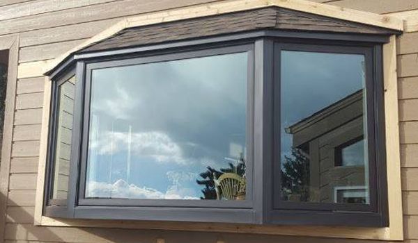 Bay window with dark frames, brown roof, reflecting a cloudy sky. Set in light brown siding.