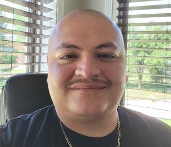 Man with shaved head and mustache smiles, wearing a black shirt, in front of a window with blinds.