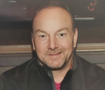 Man with light skin smiles, wearing a black jacket and a pink shirt.