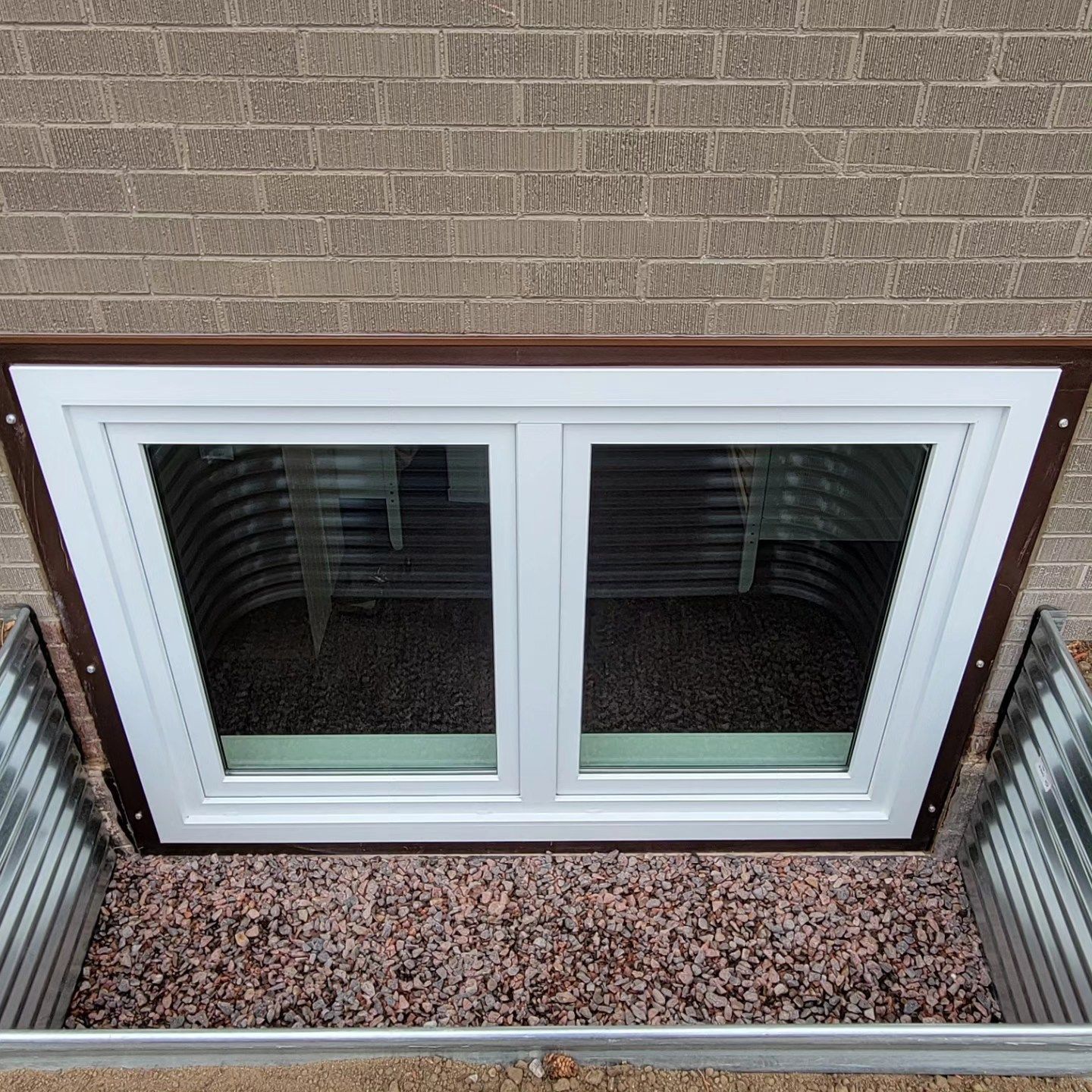 Window in a window well, surrounded by a metal and gravel, set against a brick wall.