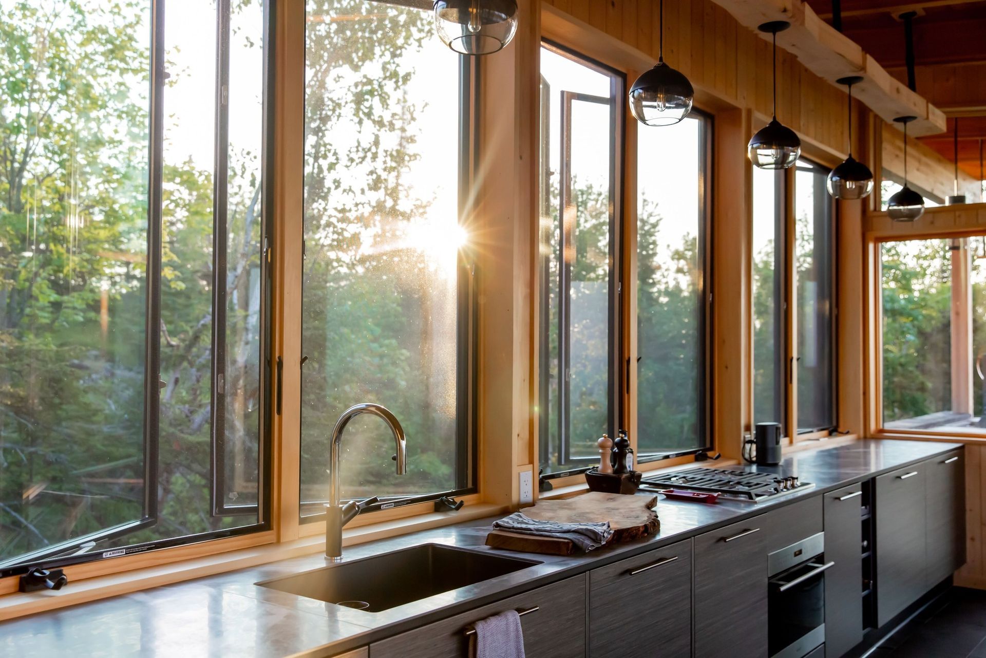 Kitchen with large windows, sunlight, sink, and countertops. Trees visible outside windows.