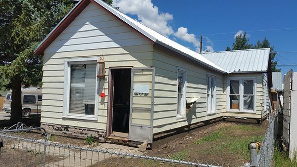 Small, weathered, one-story house with open door, light siding, and metal roof on a sunny day.