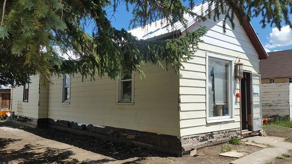 Small, off-white house with a gabled roof. Green tree branches obscure part of the view.