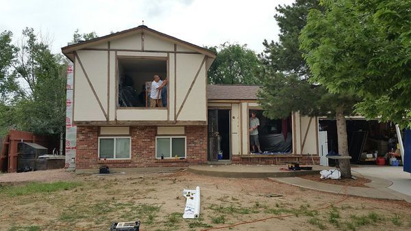 House under renovation with people in open windows, tan siding, brick, and dry lawn.