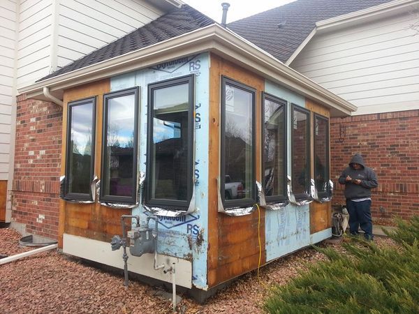 A sunroom addition under construction on the side of a brick house with visible insulation and a person standing nearby.