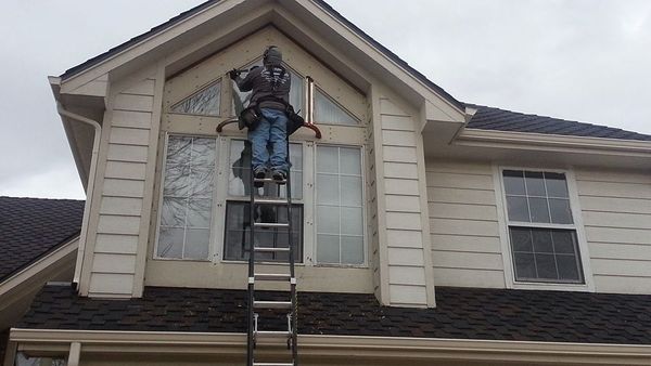 Person on a ladder cleaning windows of a two-story house with beige siding and dark roof. Cloudy day.