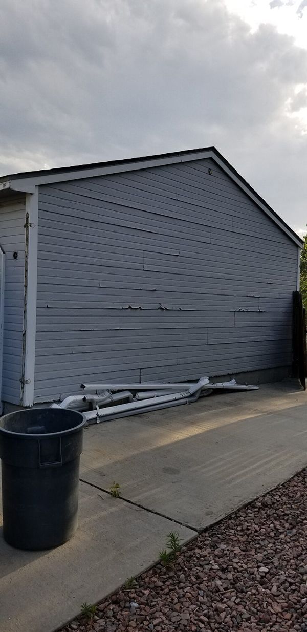 A gray building with damaged siding, a trash can, and a ladder on a concrete surface under a cloudy sky.