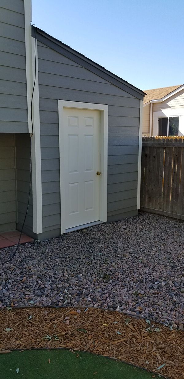 A white door on a gray building with gravel ground in front, fenced yard on right, blue sky.