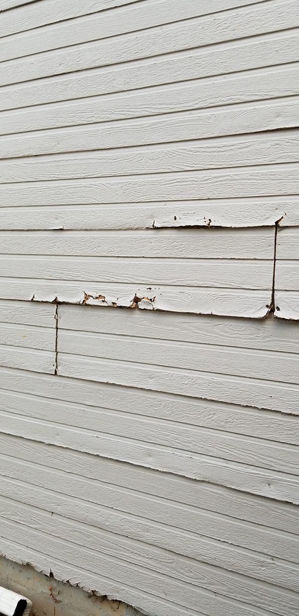 Damaged white siding on an exterior wall, with two vertical splits and missing paint.