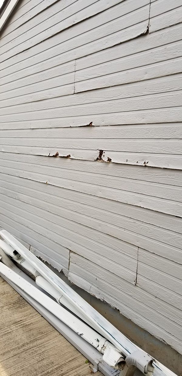 Gray siding on a building with damaged panels, possibly weathering. Ladders rest on the ground in the foreground.