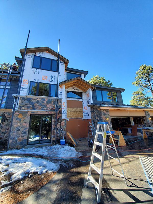 House under construction with stone and blue siding, large windows, ladder, and a clear blue sky.