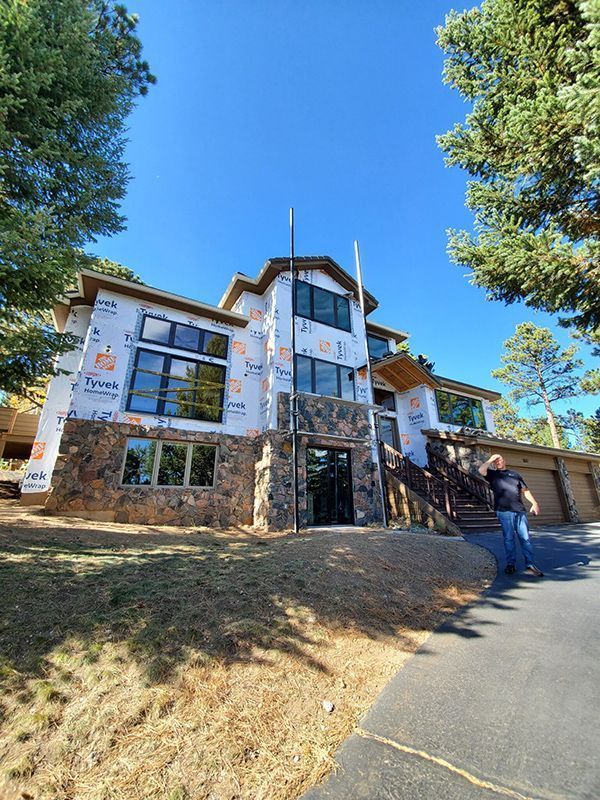 Multi-story house under construction, with stone and wrapped exterior, clear sky. Person stands near driveway.