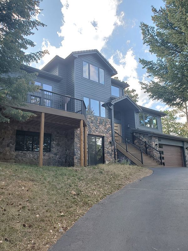 Two-story house with dark siding, stone facade, and a deck. A driveway leads to a garage. Overcast sky.