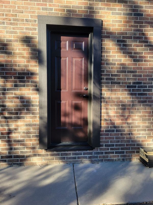 A maroon door set in a dark frame on a brick wall, with sunlight and shadows.