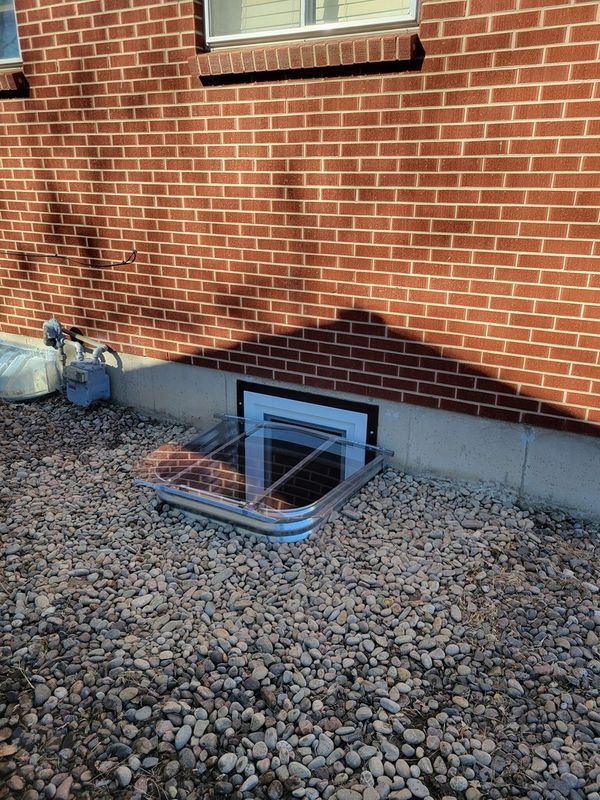 Brick building exterior with window well covered by a clear, curved lid, surrounded by gravel.