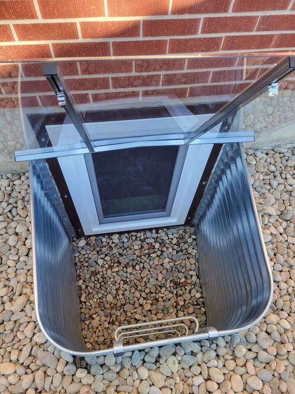 Window well with clear cover and gravel at bottom, built into a brick wall.