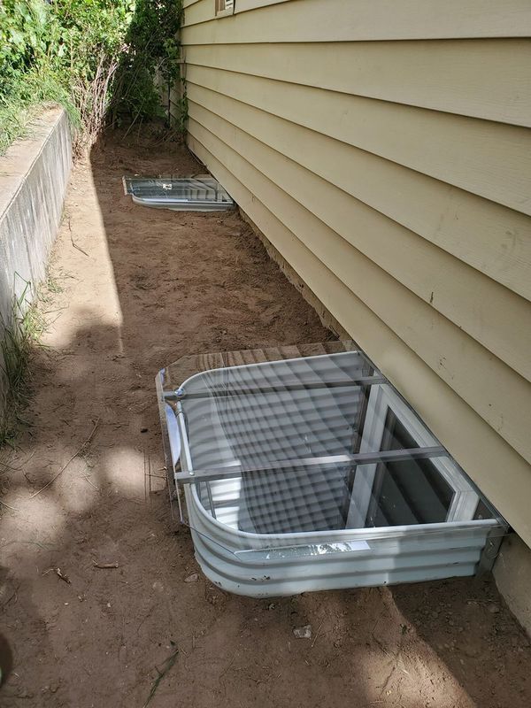 Two window wells with screens sit against a light brown house exterior. The ground is dirt.