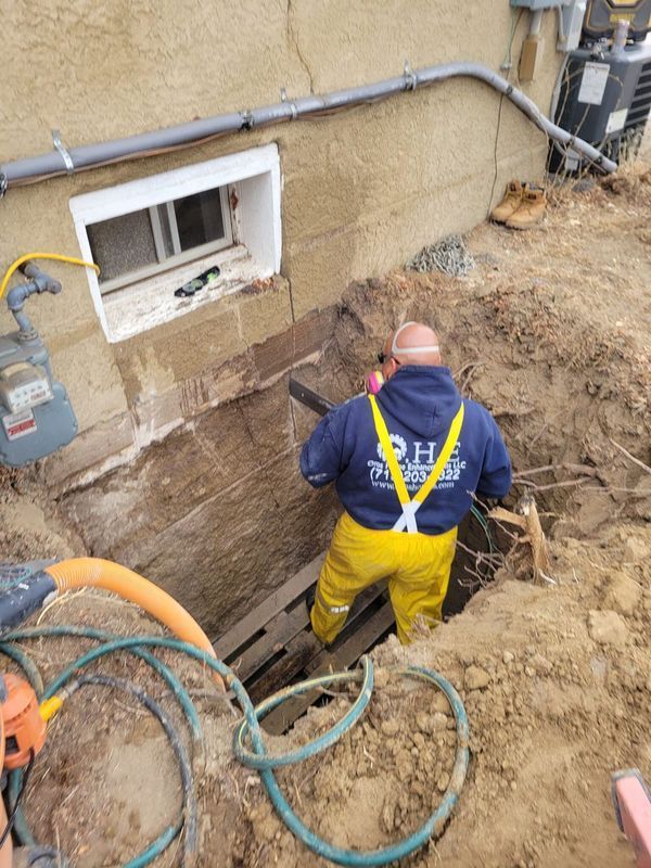 Man in work clothes uses a saw in a trench next to a building. Utility lines and window visible.