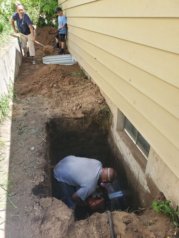 Men digging a trench next to a yellow building. One man is using a tool.