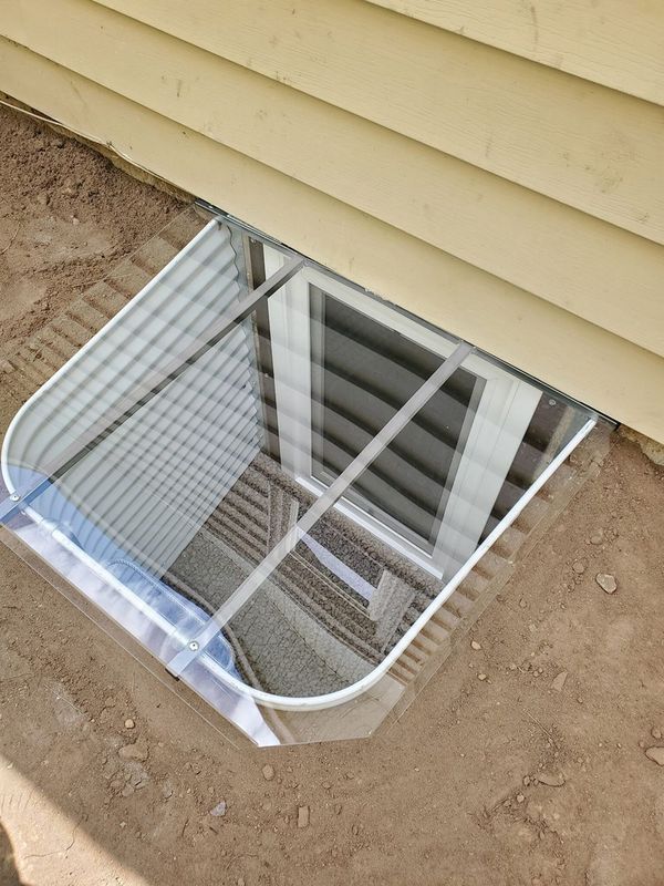 Window well with clear cover against a light yellow house. Well is metal, set in dirt.