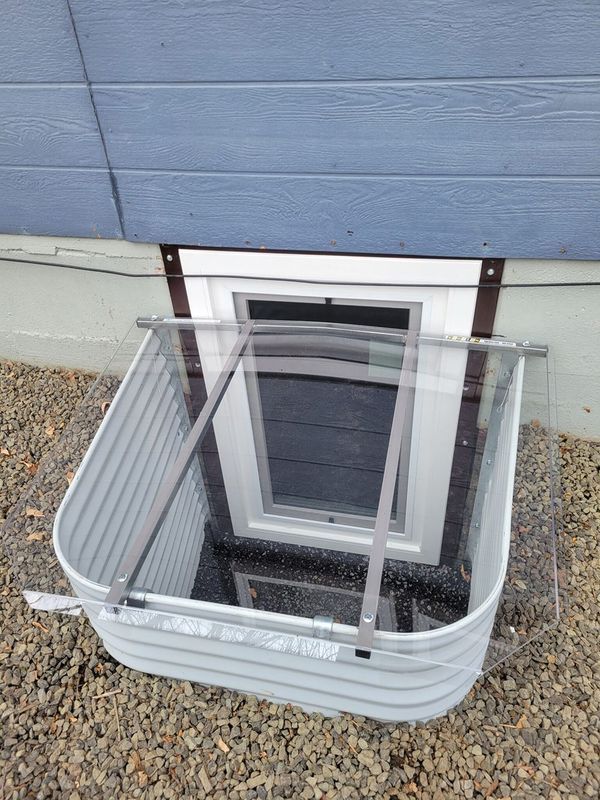 Window well with a clear plastic cover, set against blue siding.