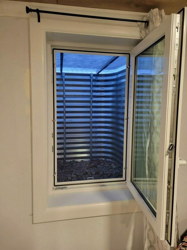 White-framed window opened, revealing a corrugated metal window well filled with rocks; blue light inside.