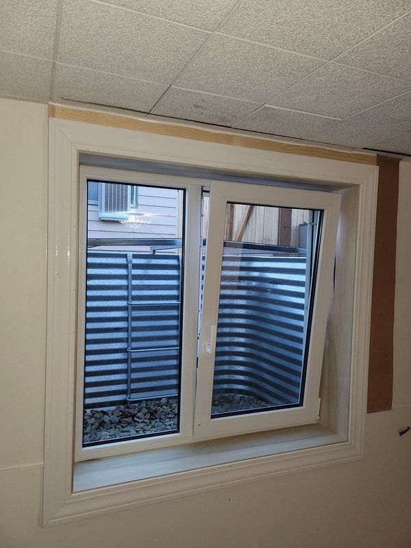 White framed basement window with one pane open, looking out at a corrugated metal window well.