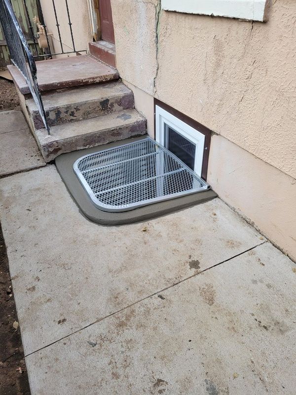 Basement window well with metal grate and concrete border, next to concrete steps and tan stucco wall.